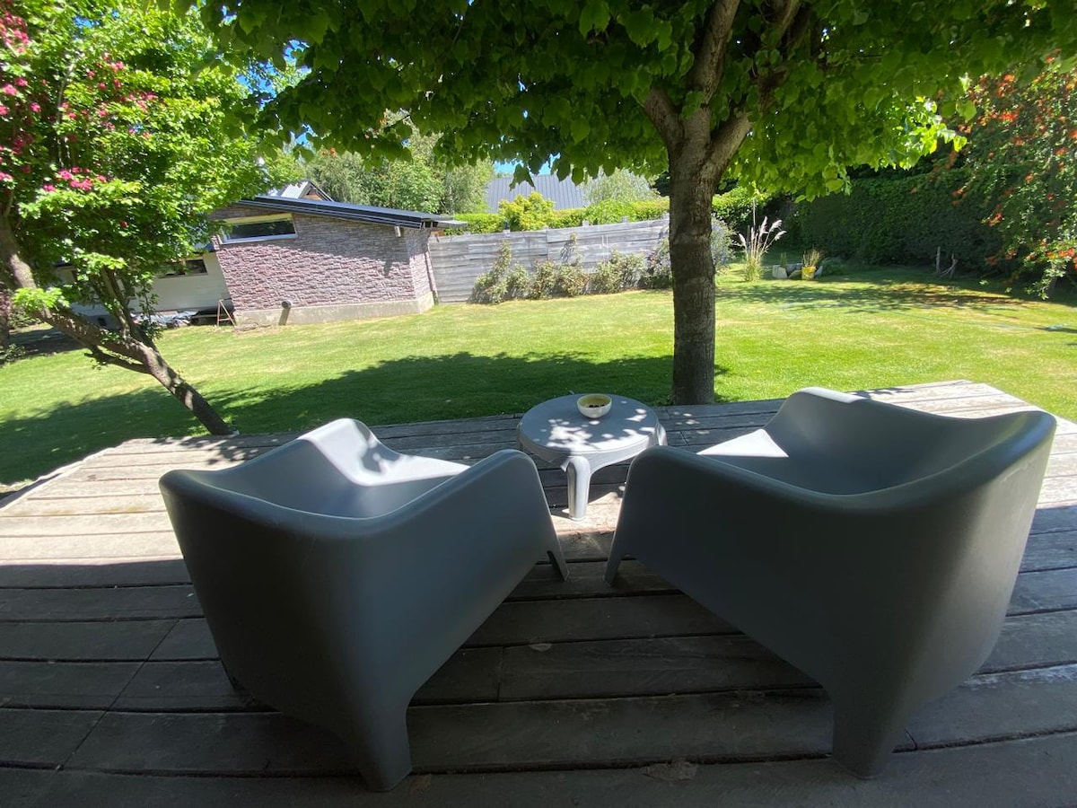 Two comfortable outdoor chairs are seen facing a small table on a wooden deck. Lush green grass and trees provide a serene backdrop, with a view of a house and garden in the distance.