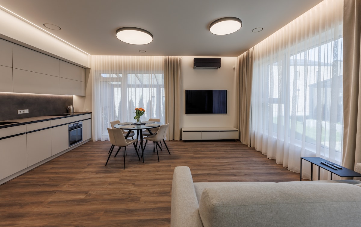 A modern living area features a light-toned sofa facing a flat-screen television mounted on the wall. A round dining table with four chairs is positioned beside expansive windows, where sheer curtains allow soft natural light to filter through, illuminating the warm wooden flooring.