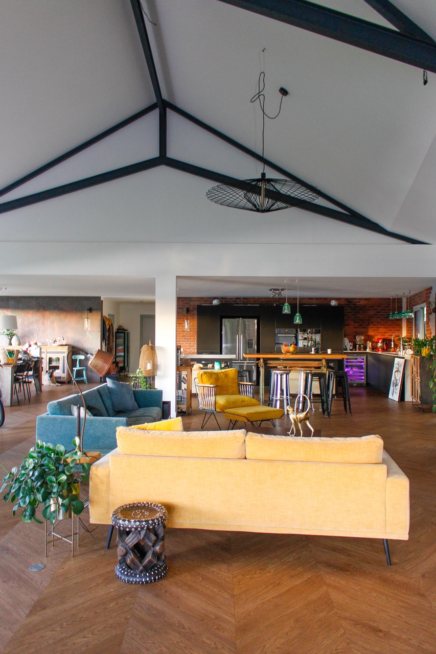 The open living area showcases a mix of colorful seating arrangements, including a yellow sofa and teal chairs. Industrial-style lighting hangs from the ceiling, while exposed beams add character. A dining area can be seen in the background, highlighting a sleek kitchen and vibrant decor.