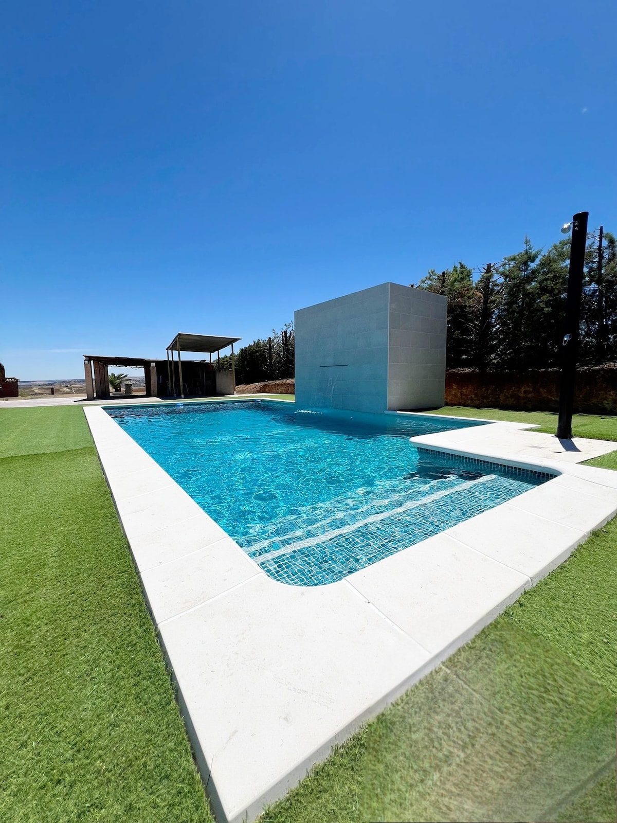 A modern swimming pool is surrounded by a well-manicured grassy area. The clear blue water reflects the sunlight, and a minimalistic white wall stands beside the pool, enhancing the open outdoor space. A shaded seating area is visible in the background.