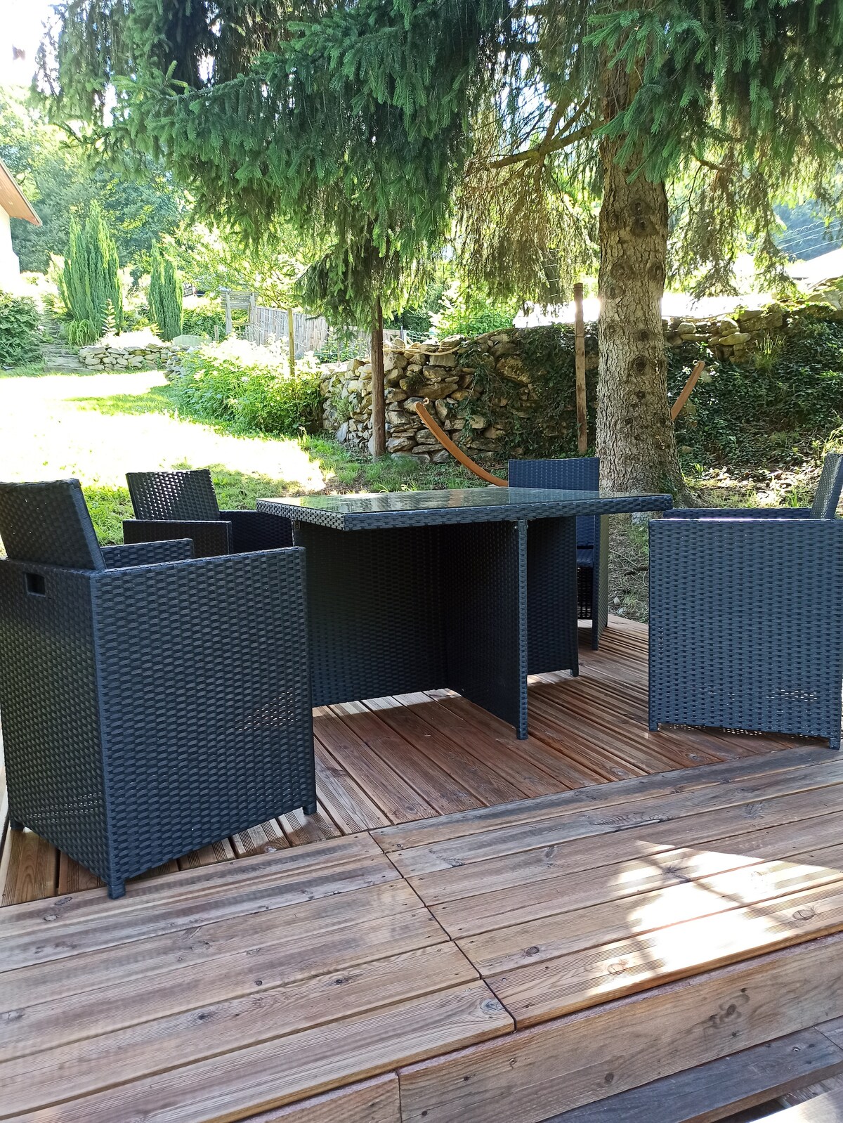 An outdoor seating area is presented, featuring a rectangular table surrounded by four rattan chairs. The setting is shaded by a large tree, with sunlight filtering through the branches, illuminating the wooden deck beneath.