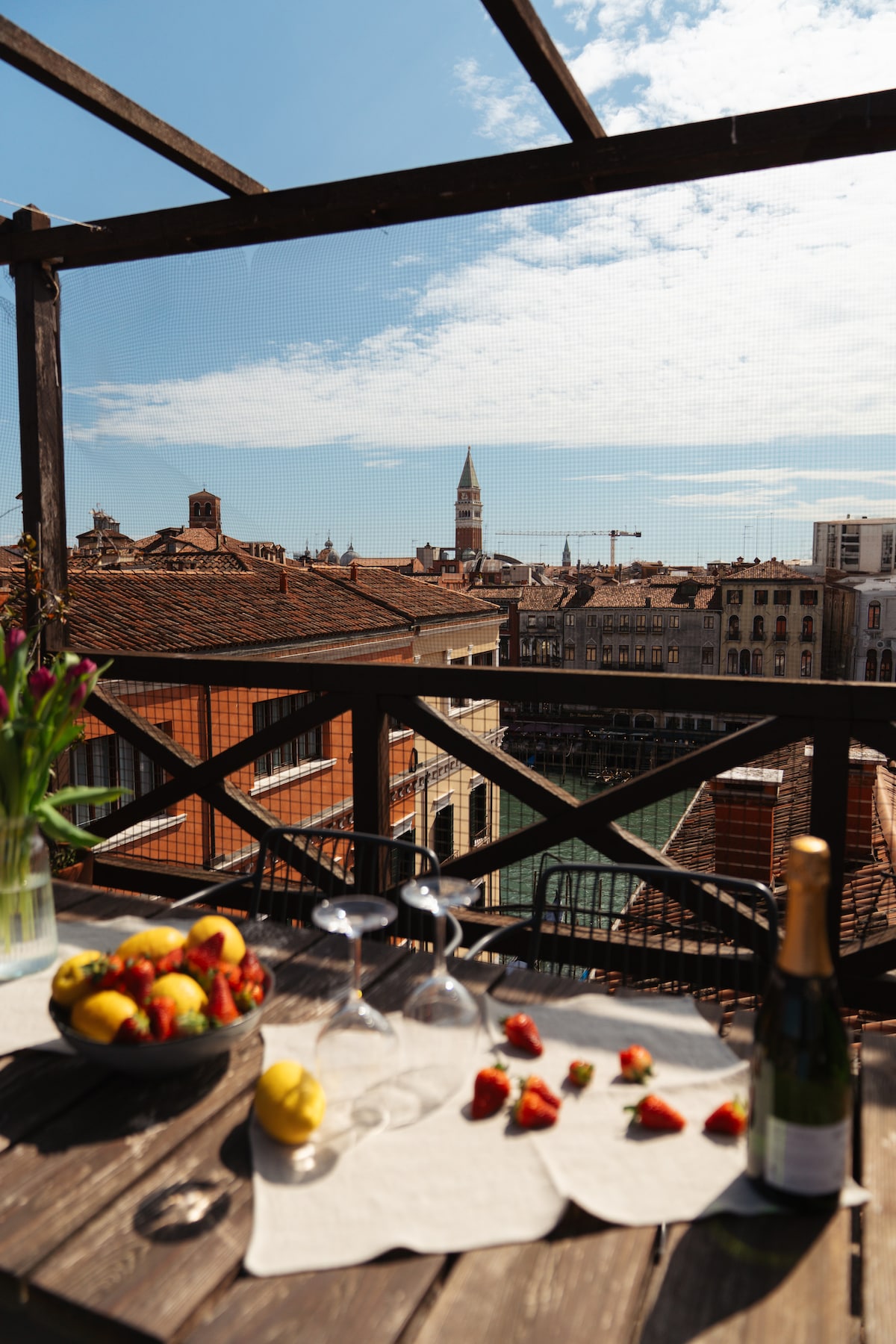 A rooftop terrace features a wooden table adorned with fruit, glasses, and a bottle of sparkling drink. A scenic view of Venetian rooftops and a bell tower is visible in the distance, complemented by a clear blue sky with scattered clouds.