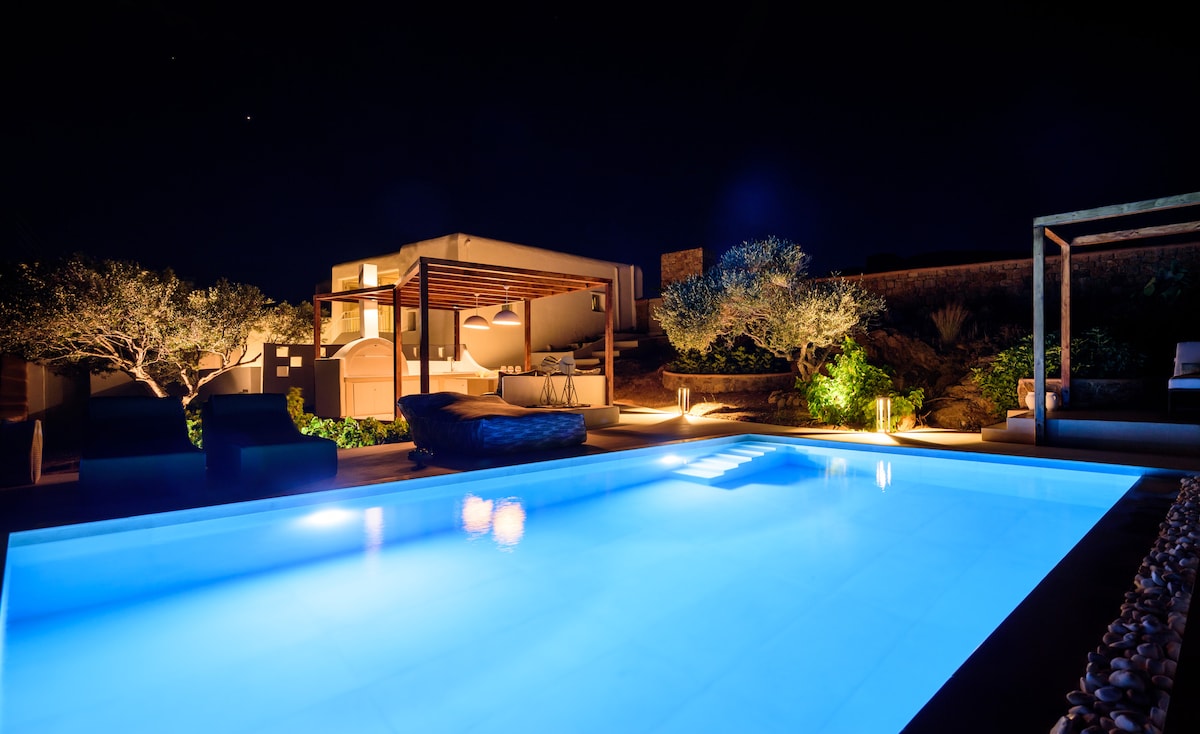 The private pool is softly illuminated, reflecting a serene blue hue against the dark surroundings. Comfortable outdoor seating can be seen nearby, along with illuminated greenery, creating a calm and peaceful outdoor space.