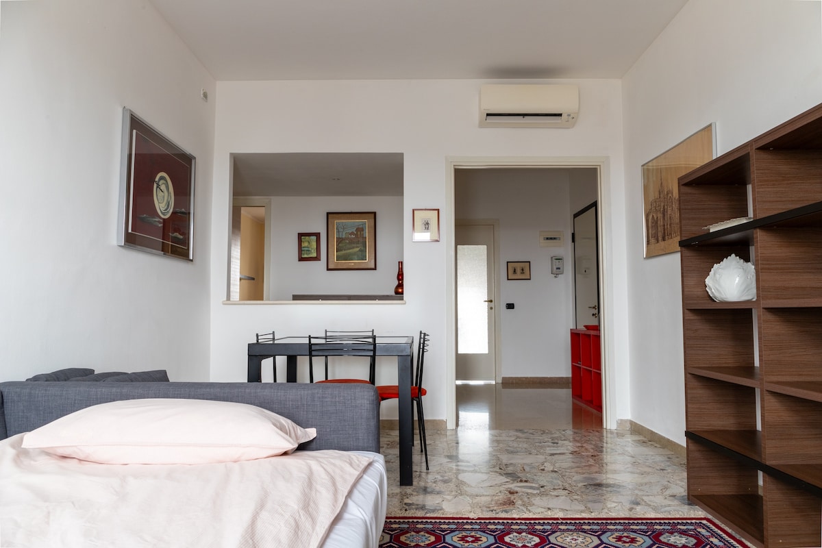 The living area features a comfortable sofa bed with soft bedding. A minimalist dining table accommodates four, set against light-colored walls adorned with framed artwork. A stylish bookshelf is positioned in the corner, while the entrance leads to a well-lit hallway.