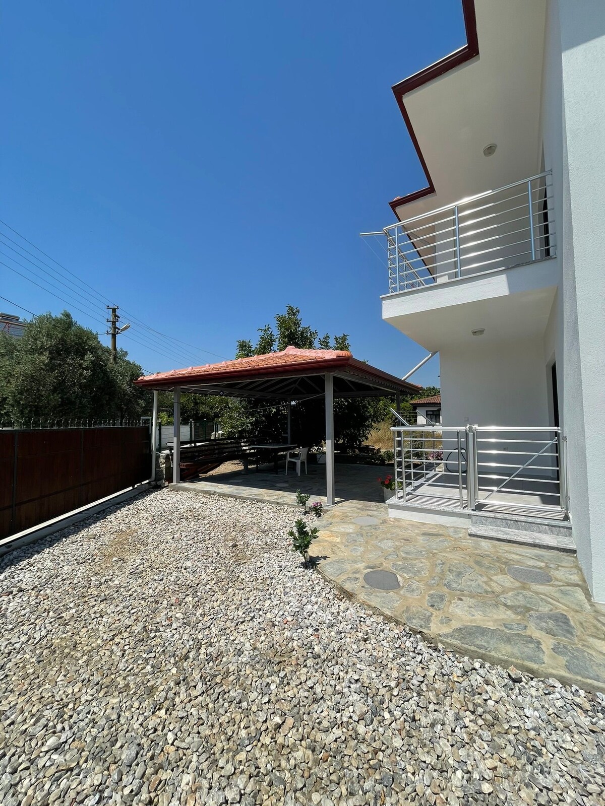 A well-maintained outdoor space is featured, with a covered patio area supported by metal columns. Pebbled ground adds texture, while greenery surrounds the space. The building's modern architecture is complemented by a balcony on the upper level.