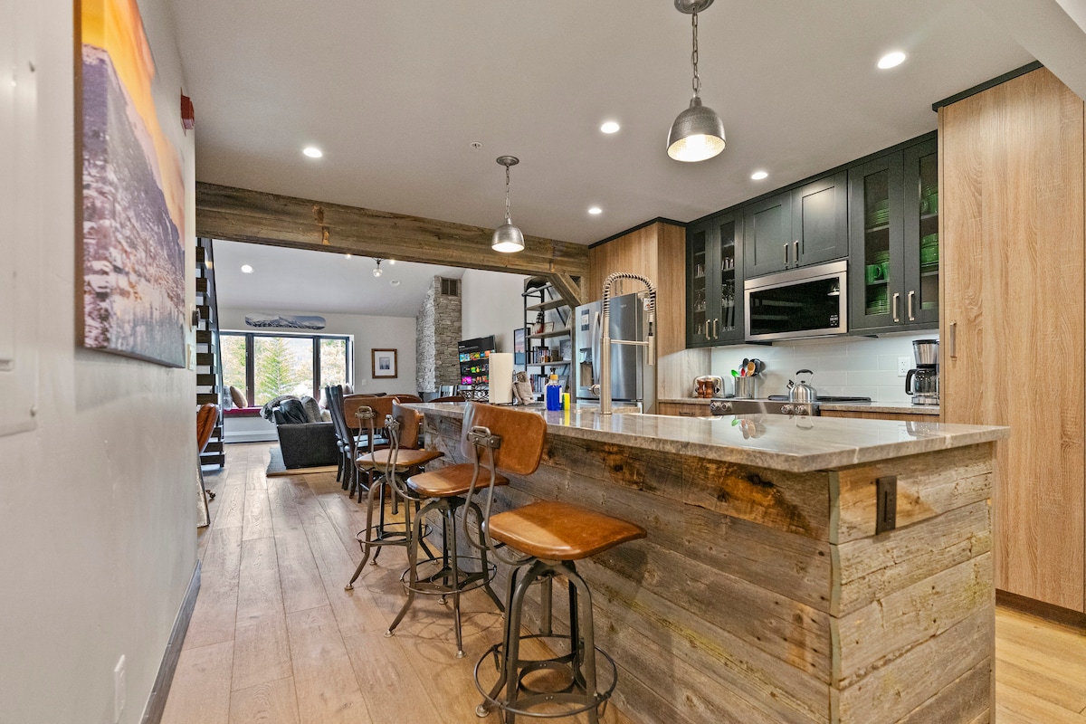 An open-concept kitchen features a large granite island with four wooden bar stools. Dark cabinetry and stainless-steel appliances create a modern space. Natural light fills the area from large windows, illuminating the cozy living room beyond, which is furnished with plush seating.