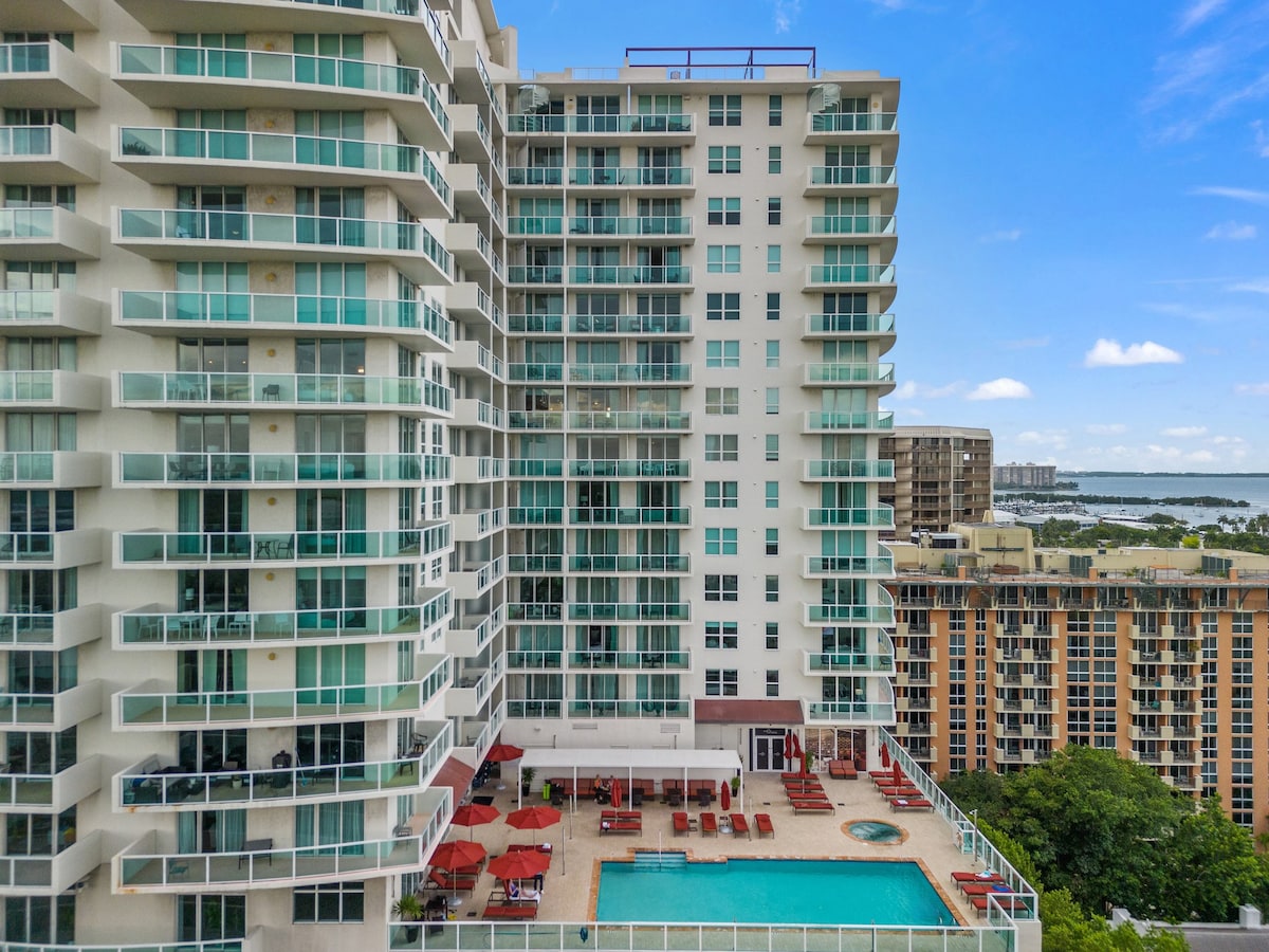 The exterior of a modern 22-story building is depicted, featuring a sleek design with numerous balconies. A pool area with red lounge chairs is visible in the foreground, surrounded by a glass railing. The backdrop showcases vibrant city views and a glimpse of the bay.