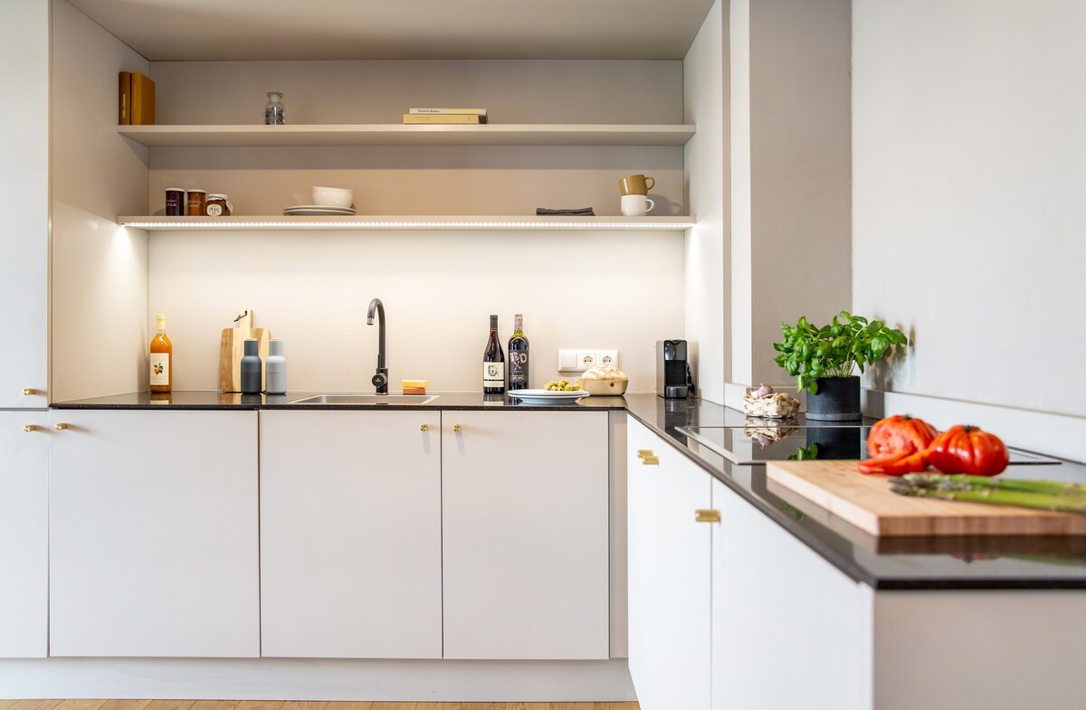 A modern kitchen is presented with sleek white cabinetry and a dark countertop. Open shelves display various items, including bottles and tableware. A cutting board with fresh tomatoes is visible, alongside a stainless-steel sink and coffee machine, enhancing the practical design.
