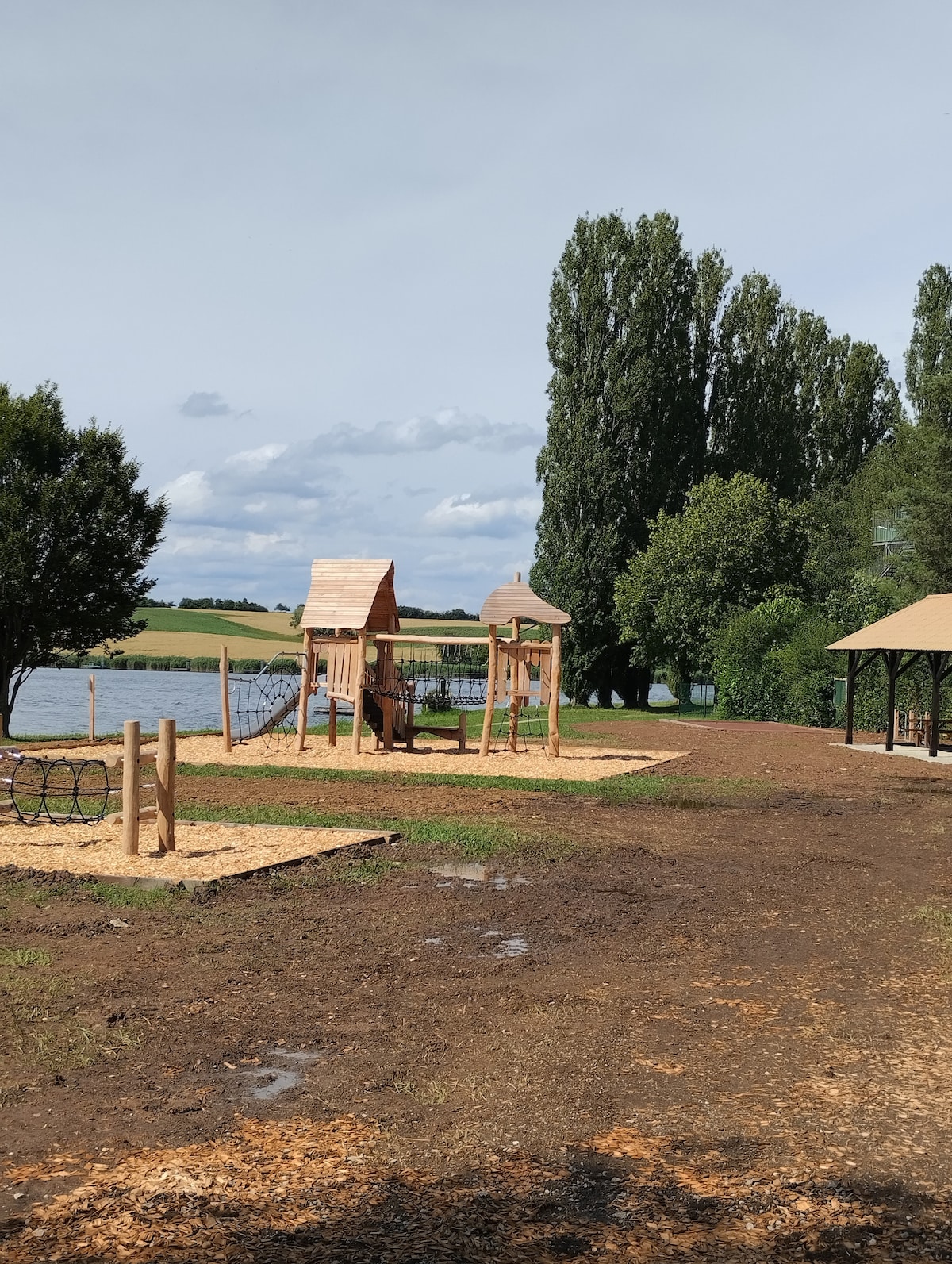 A recreational area is visible alongside a serene pond, featuring wooden play structures designed for children. Soft mulch covers the ground, enhancing safety. Nearby, trees provide shade, and open green spaces are present, inviting outdoor activities in a peaceful environment.