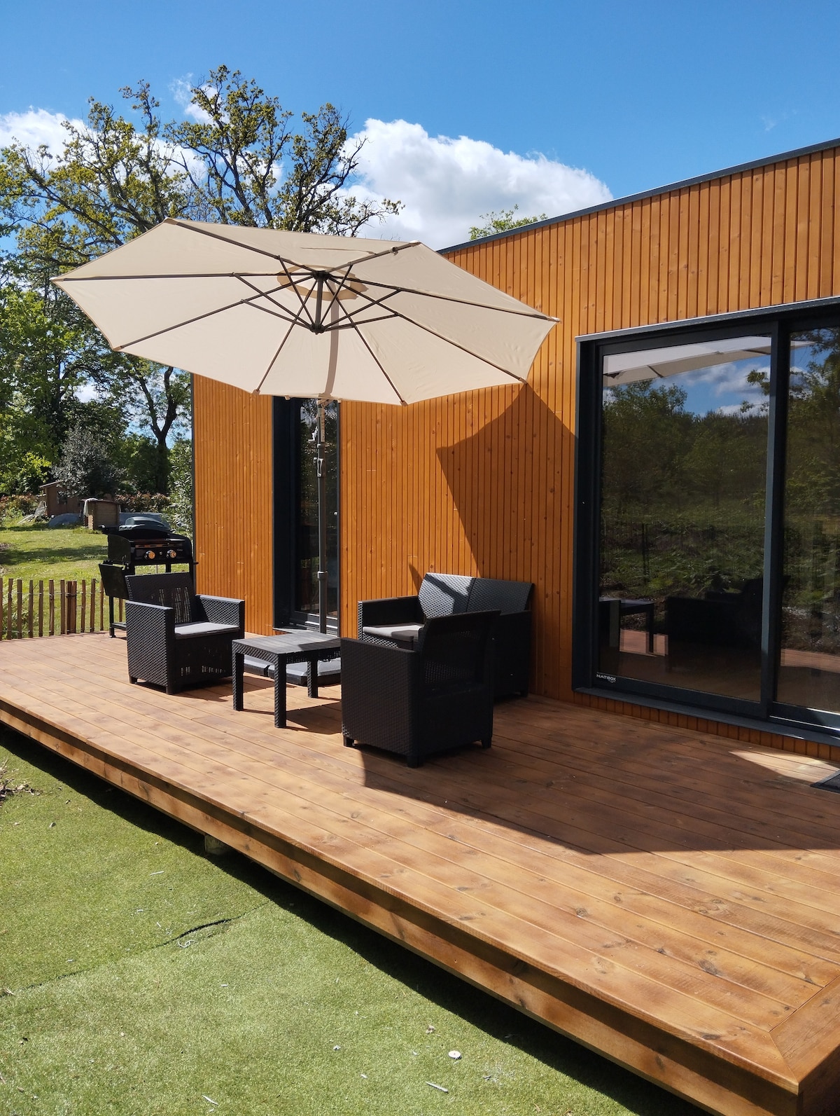 A wooden terrace features a light-colored parasol providing shade over a set of patio furniture, including two armchairs and a small table. The backdrop includes a wooden exterior of the accommodation, with greenery visible in the surrounding area.