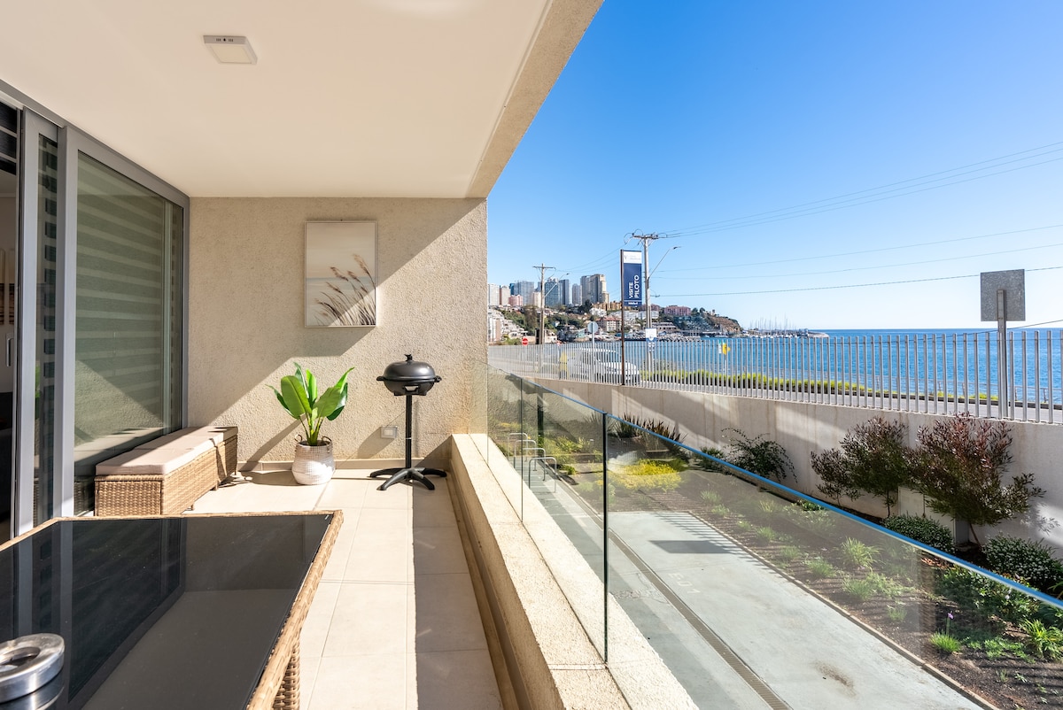 A spacious terrace features a glass railing, providing an unobstructed view of the coastline. A grill is positioned on one side, and a potted plant adds a touch of greenery. The area is complemented by a comfortable lounge chair, enhancing the outdoor ambiance.