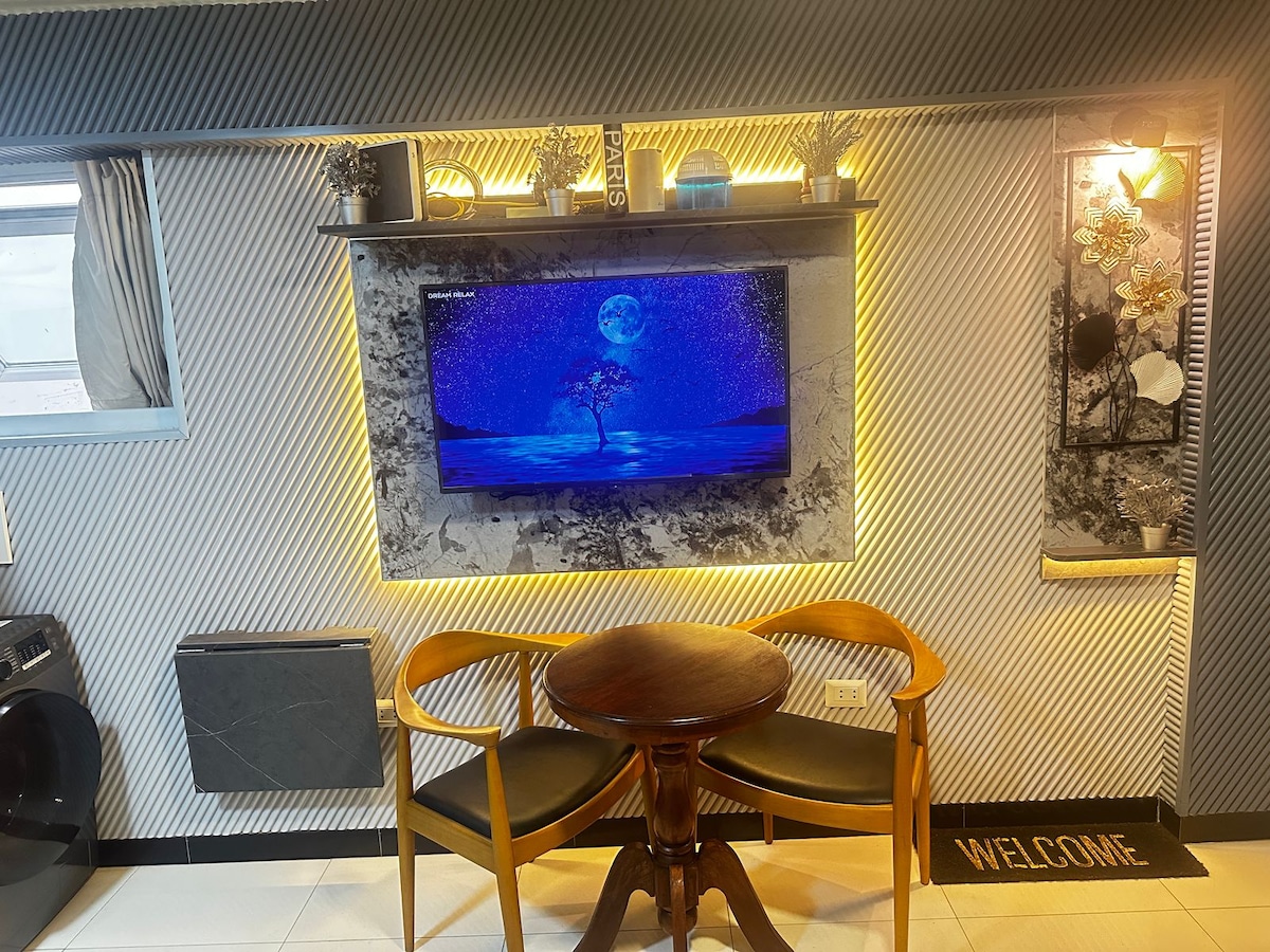 A small dining area features a round wooden table accompanied by two black chairs. Above, a wall-mounted Smart TV displays a serene nighttime scene. Decorative plants adorn the shelves, while ambient lighting enhances the overall aesthetic.