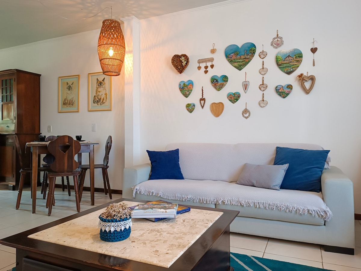 A cozy living area features a light-colored sofa adorned with blue cushions, complemented by a coffee table that holds decorative items. A wall display of various heart-shaped ornaments and artwork adds interest, while a dining set is visible in the background, illuminated by a woven pendant light.