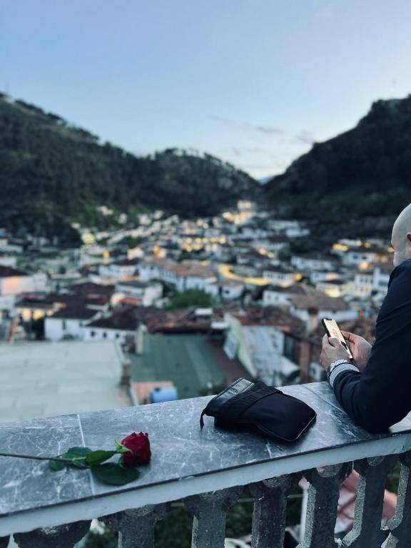A person is seen sitting on a stone railing, absorbed in a phone, overlooking a vast cityscape with a backdrop of green hills. A single red rose rests on the railing beside a dark bag, adding a thoughtful element to the serene view.