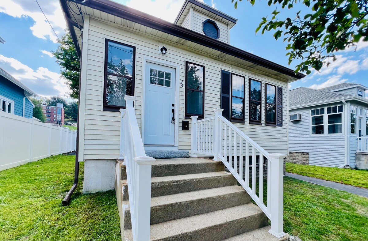 The exterior of the bungalow features a welcoming front entrance with white railings leading up to the door. Sunlight brightens the facade, highlighting the well-maintained siding and the large windows. A lush green lawn surrounds the base of the house, contributing to the inviting atmosphere.