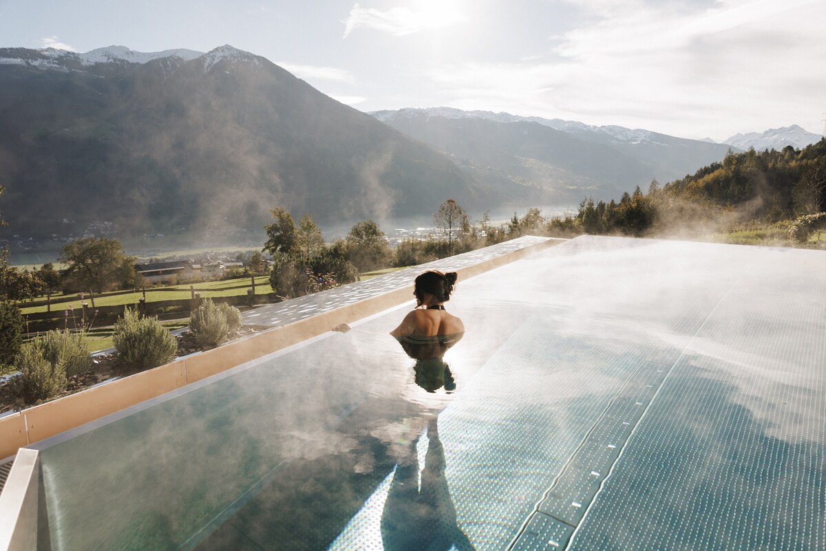A serene infinity pool stretches towards the horizon, surrounded by mist and showcasing stunning mountain views. The water reflects the sunlight, creating a tranquil atmosphere. Lush greenery lines the edge of the property, enhancing the peaceful retreat.