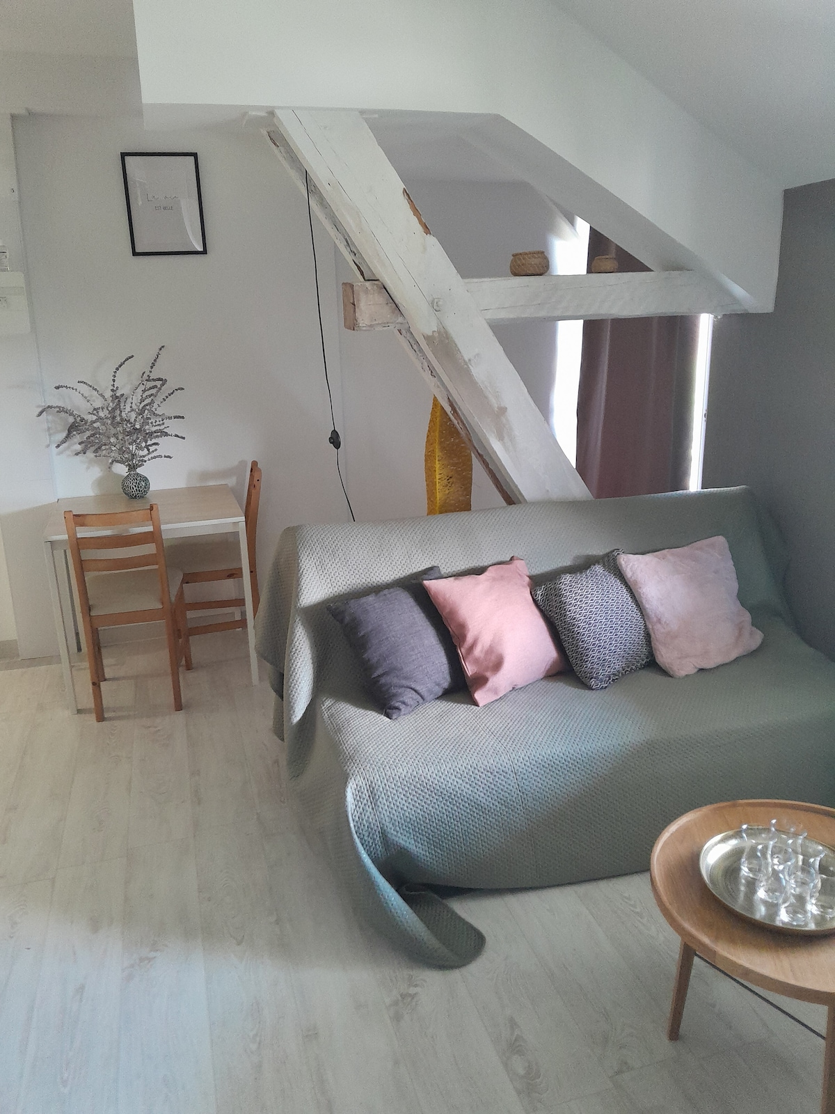A cozy corner features a sofa draped with a soft cover, adorned with several pillows in muted tones. A small dining table with two chairs is positioned nearby, complemented by light-colored wooden flooring and a blend of natural light from the window.