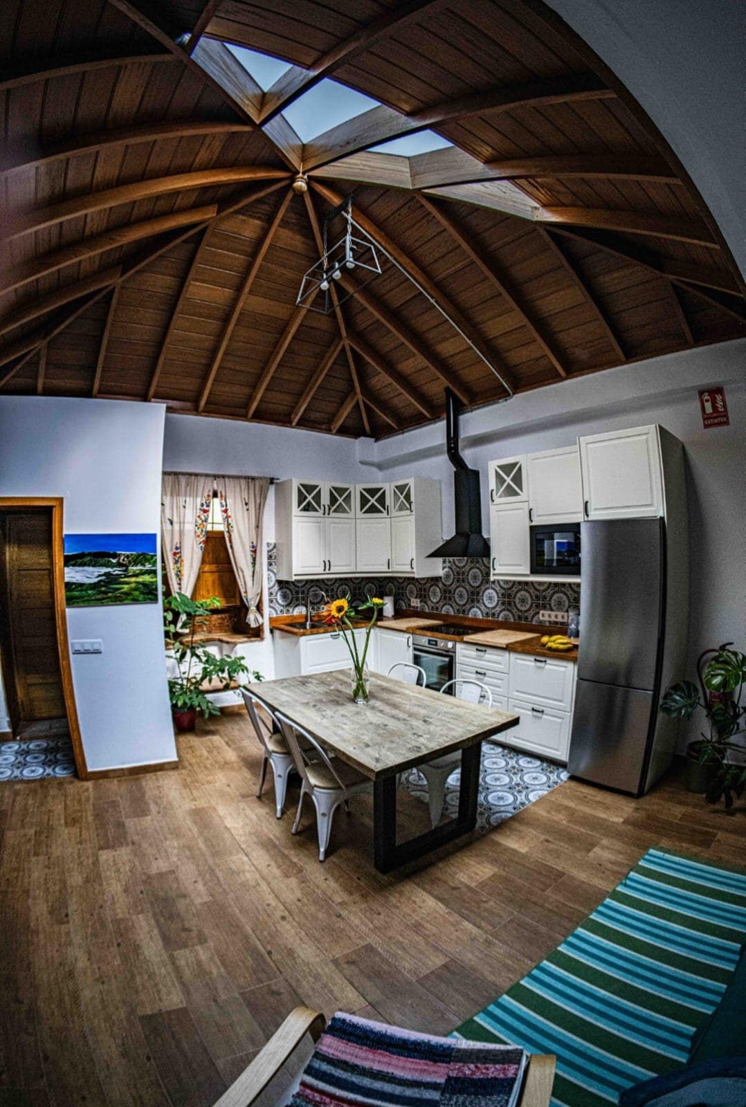 An open-concept kitchen and dining area features a triangular wood-beamed ceiling and a large skylight. A wooden dining table with light-colored chairs is complemented by decorative plants. The kitchen showcases modern appliances and tiled accents, with natural light illuminating the space.
