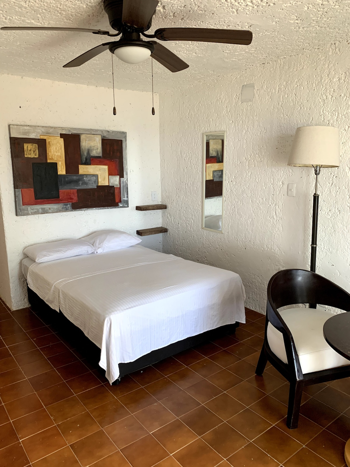 A simple room is displayed with a spacious bed made with white linens. A fan hangs from the ceiling, and a wall art piece features geometric designs in muted colors. A full-length mirror is mounted beside the bed, with a small table and chair positioned nearby.