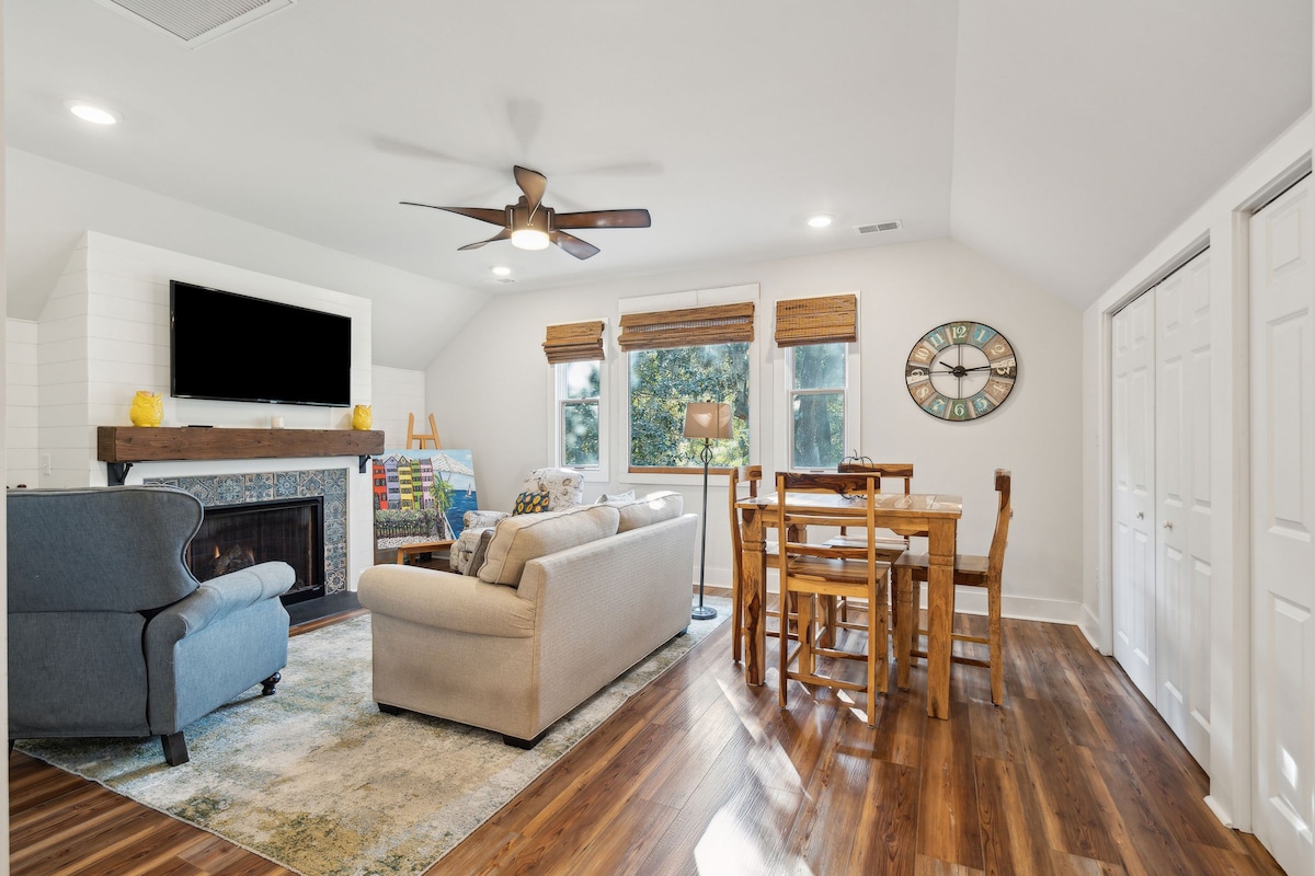 The spacious living area features a comfortable sofa and armchair, with light streaming through large windows. A dining table for four lies adjacent, and a fireplace is framed by a decorative mantel. A flat-screen TV is mounted on the wall, complemented by wooden flooring.