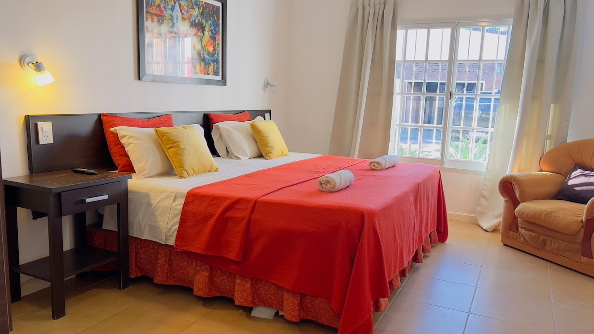 A spacious sleeping area is presented with a king-sized bed covered in a red and white bedspread. Neutral-colored pillows and rolled towels are arranged on the bed. A comfortable armchair complements the space, while large windows allow natural light to enter.