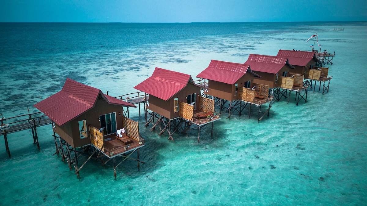 A series of water chalets are depicted on stilts above turquoise waters, featuring red roofs and wooden balconies. The serene environment is highlighted by the vast ocean, with a clear horizon and gentle waves. The chalets are positioned in a linear arrangement, enhancing the tranquil retreat atmosphere.