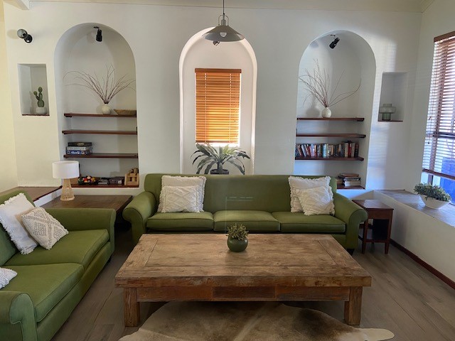 A cozy lounge area features a large, rustic coffee table at the center, surrounded by green couches with decorative cushions. Shelving units built into the wall display plants and books, while natural light filters through wooden blinds.