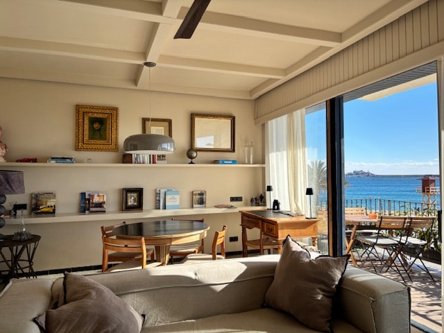 A spacious living area features a comfortable sofa adjacent to a circular dining table. Shelves hold books and decor, while large glass doors offer a view of the sea and a terrace outside. Soft natural light illuminates the room, creating an open and airy ambiance.