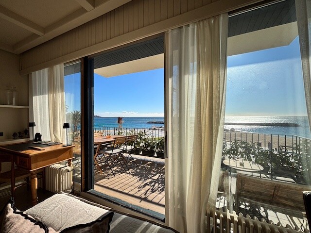 A spacious room features large glass doors that open to a balcony overlooking the sea. Soft curtains frame the view, allowing natural light to fill the space. A wooden desk is positioned near the window, providing a cozy workspace.