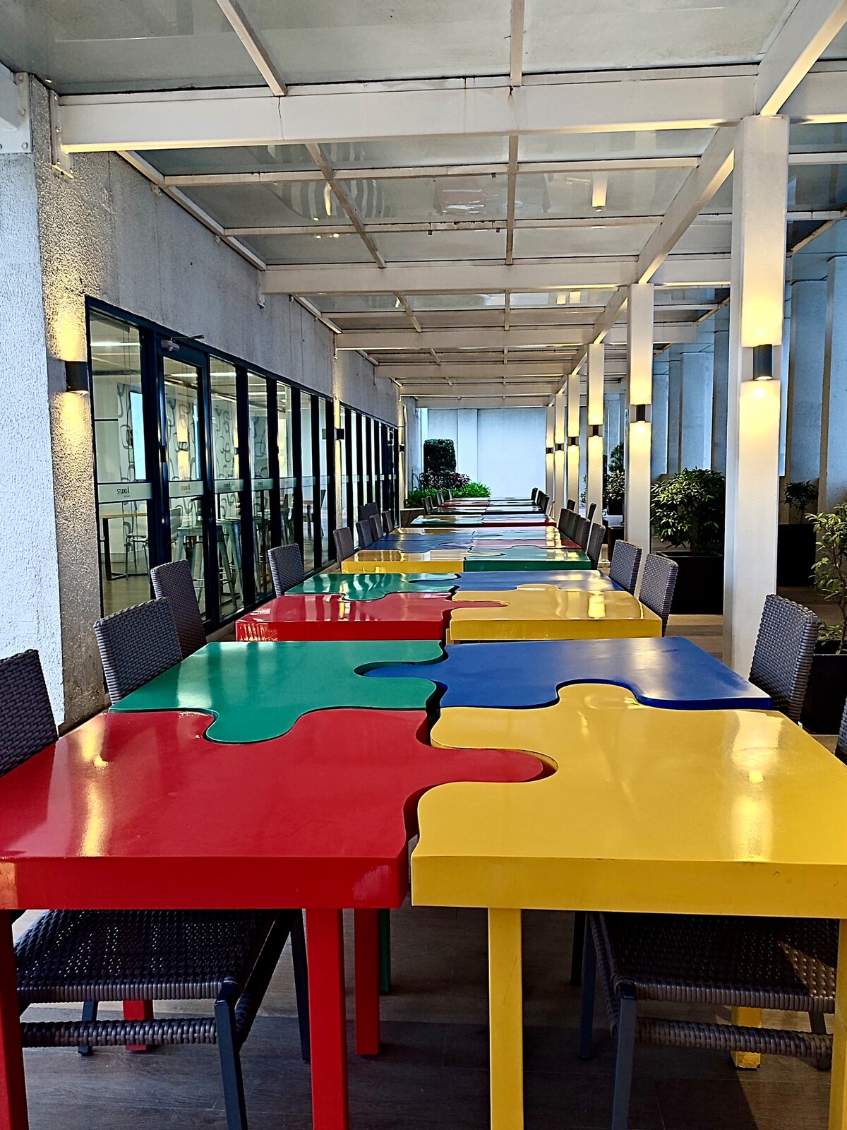 A row of colorful puzzle piece tables is displayed in an outdoor area, with vibrant red, yellow, blue, and green surfaces. Each table is accompanied by dark wicker chairs. Large windows allow natural light to filter in, framing the space with greenery and enhancing the lively atmosphere.