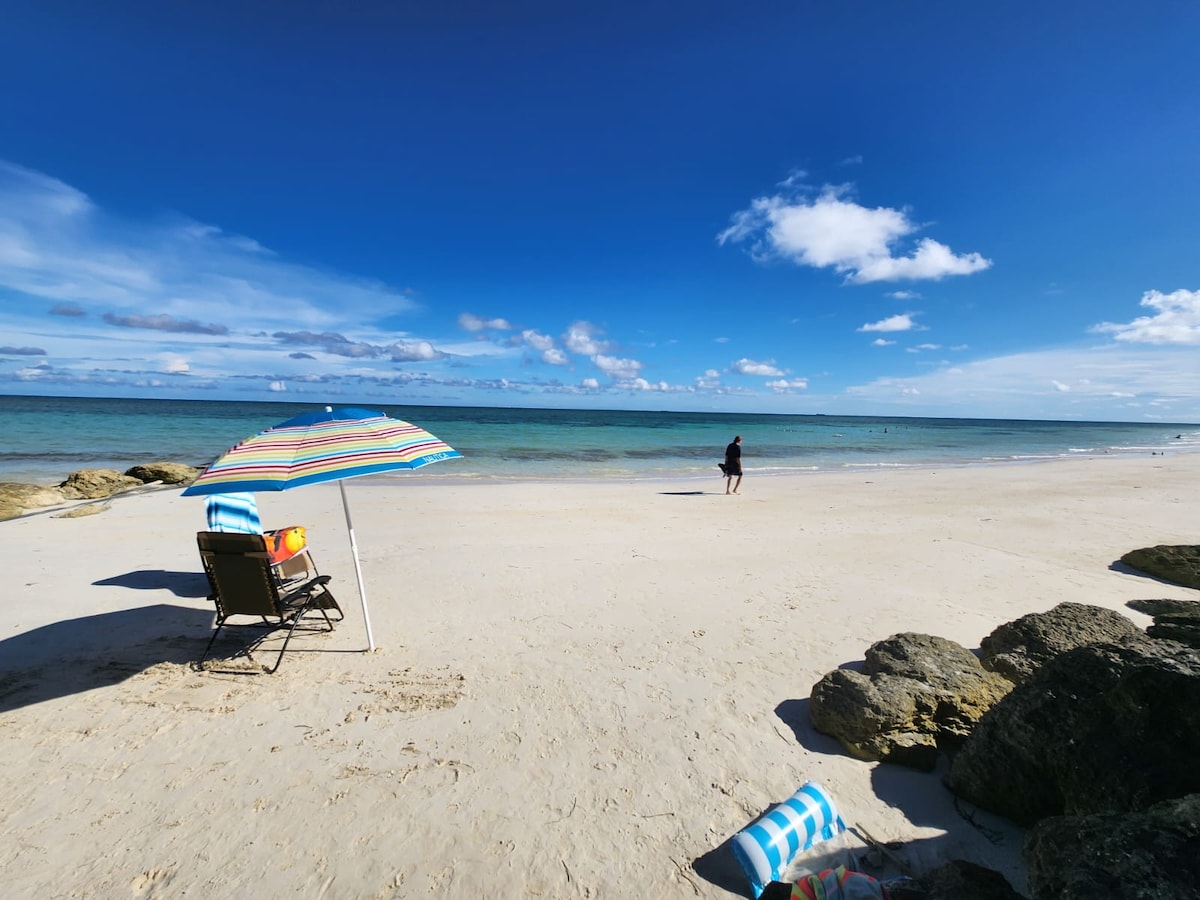A serene beach scene displays soft, sandy shores meeting clear, turquoise waters under a bright blue sky. A colorful umbrella provides shade over a seating area, while a lone figure strolls along the water's edge. Nearby, beach towels rest on the sand.