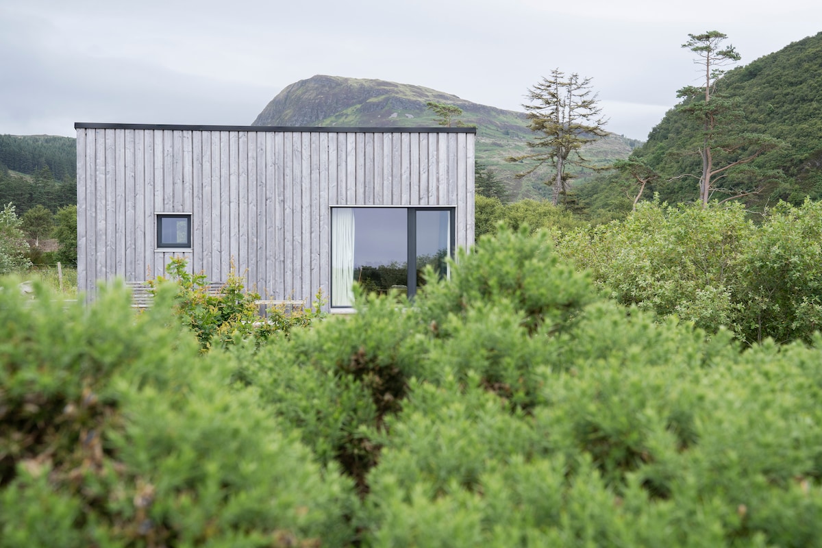 A modern cabin is nestled among green foliage, displaying a natural wooden exterior. Large windows reflect the surrounding landscape, and a gentle slope of hills is visible in the background, enhancing the sense of secluded tranquility.