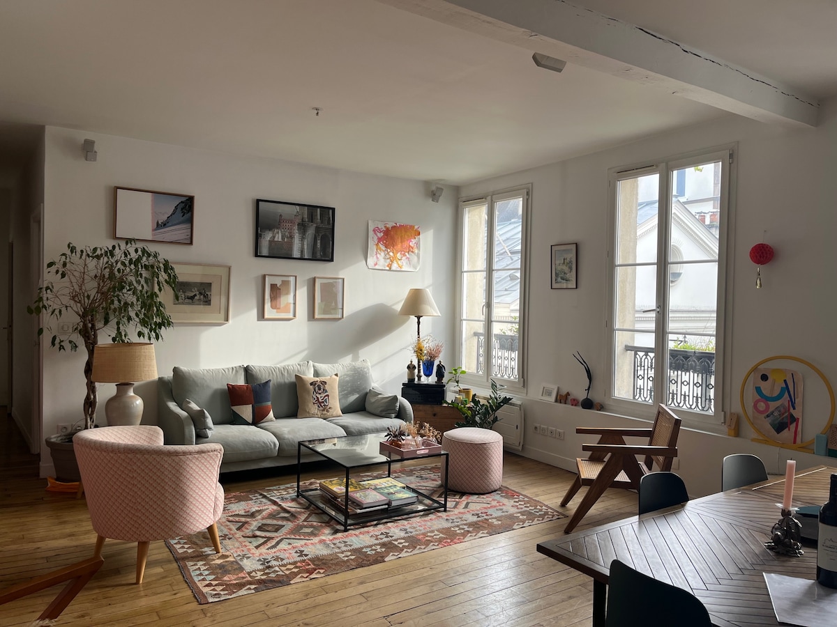 A spacious living area is bathed in natural light from large windows. Cozy seating is arranged around a coffee table with decorative items. Wall art enhances the ambiance, while a dining table occupies one corner. Wooden flooring adds warmth to the overall design.