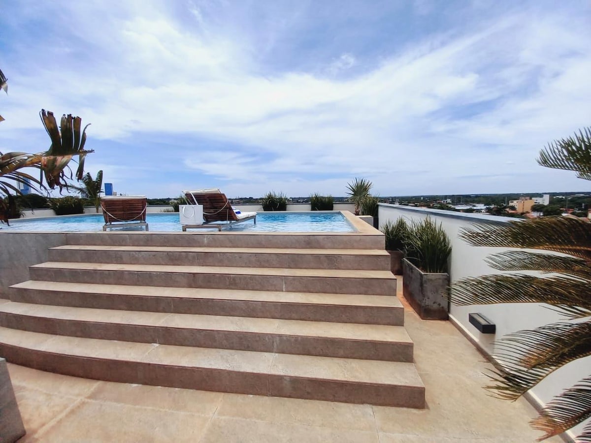 A rooftop terrace is featured with a stunning swimming pool, creating a serene environment for relaxation. Two lounge chairs are positioned along the edge, offering views of the city skyline. Surrounding greenery enhances the tranquil setting, complemented by expansive steps leading down from the pool area.