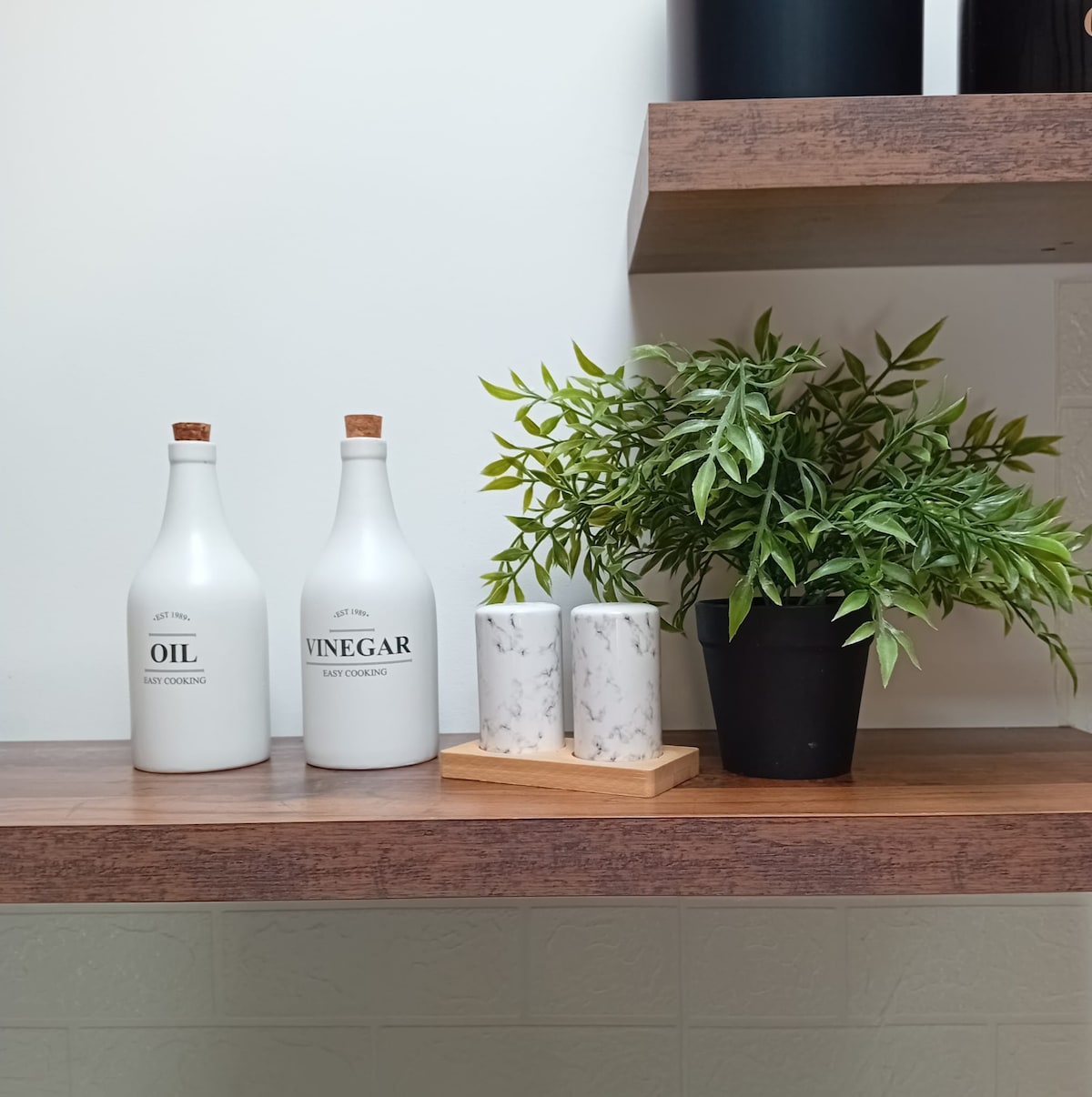 On a wooden shelf, two white bottles labeled 'OIL' and 'VINEGAR' are presented alongside a small black pot containing a green leafy plant. Also visible is a rectangular wooden tray holding two white marble-patterned candles, enhancing the kitchen decor.