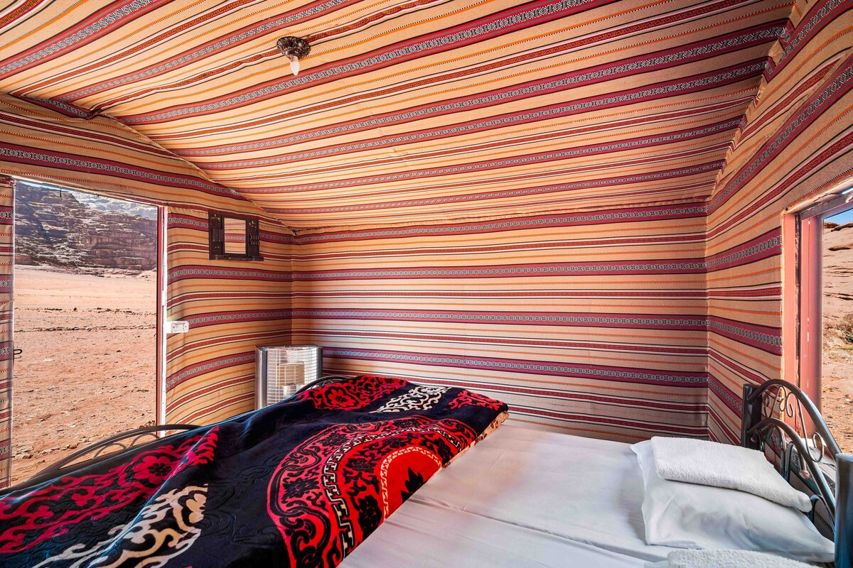 A tent interior is adorned with colorful, striped fabric, creating a warm ambiance. A spacious bed is dressed with a patterned blanket and a set of pillows. Natural light enters through an open door, offering a view of the surrounding landscape.