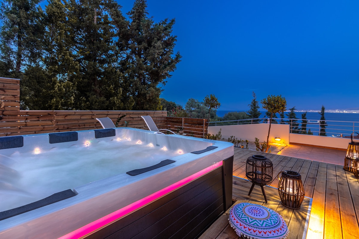 A private jacuzzi is nestled on a wooden terrace, surrounded by lush greenery under a twilight sky. Soft ambient lighting enhances the inviting atmosphere. Two lounge chairs and stylish lanterns complement the serene setting, offering a tranquil retreat with scenic views of the surroundings.