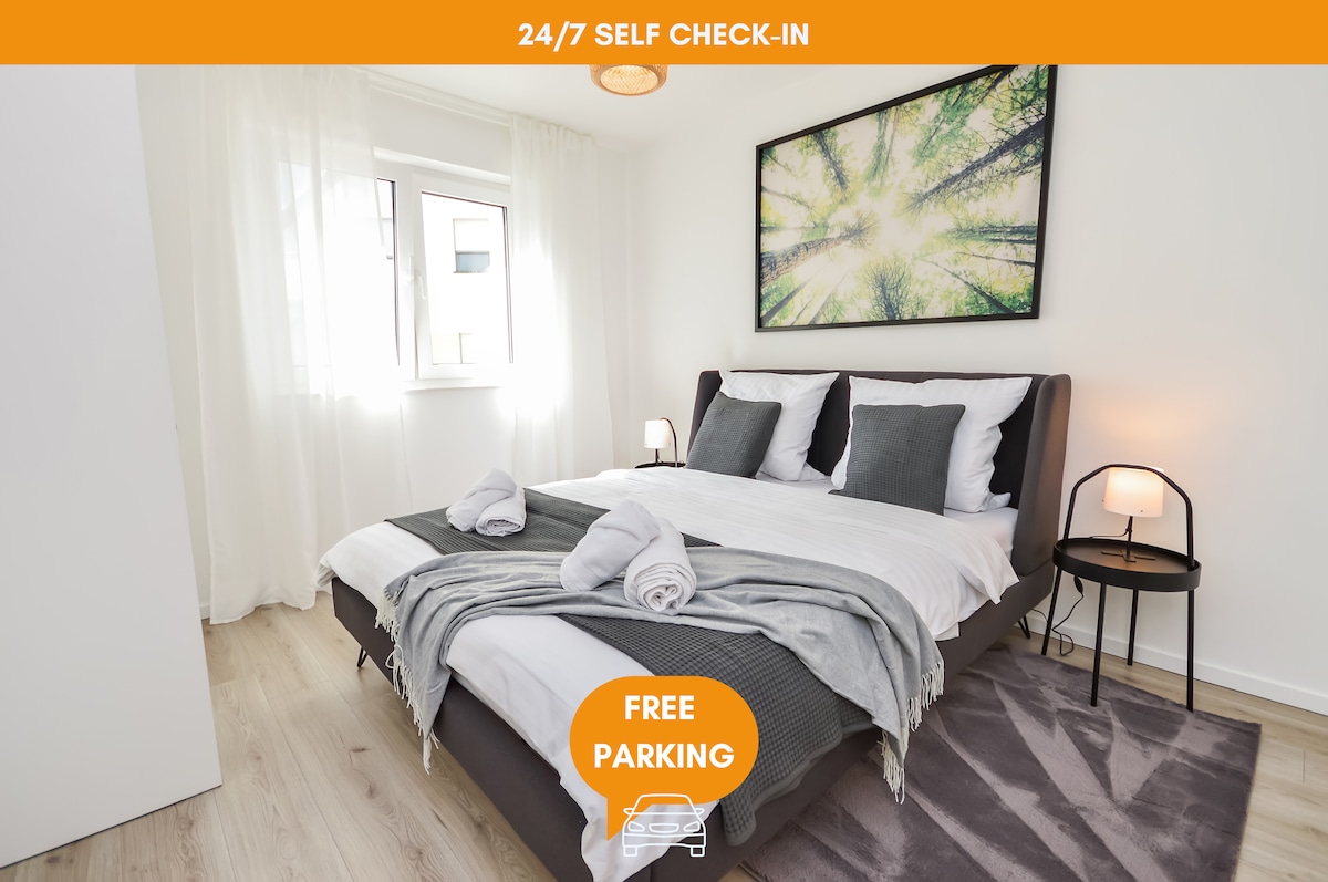 A bright bedroom features a spacious bed dressed in neutral linens and decorative pillows. Natural light filters through sheer curtains, illuminating the wood flooring. A large piece of wall art depicting a forest scene enhances the calm ambiance. A bedside table with a lamp completes the space.