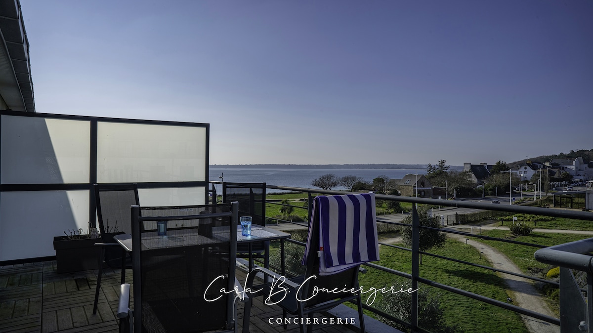 A balcony equipped with outdoor furniture offers a view of the sea and surrounding landscape. A striped towel hangs over the chair, and the serene blue sky complements the tranquil atmosphere. The area is surrounded by greenery and a pathway, enhancing the peaceful setting.