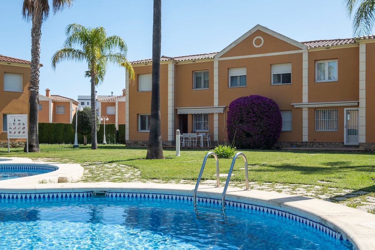 The image showcases a well-maintained property with a bright orange facade, surrounded by palm trees and lush greenery. A swimming pool is visible in the foreground, featuring clear blue water and a set of steps leading in. Tables and chairs can be seen on the patio area.