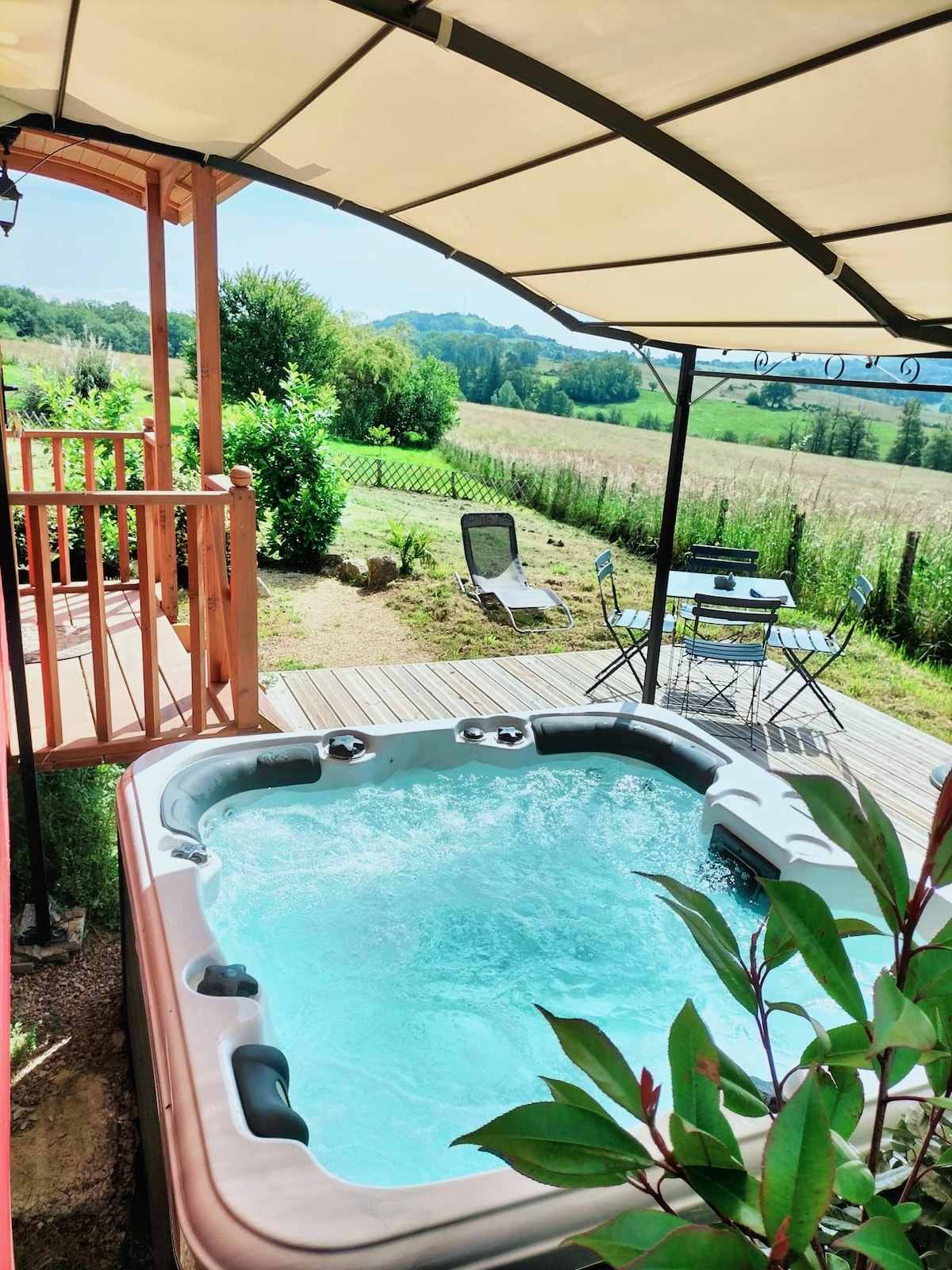 A private, covered hot tub is positioned on a wooden terrace, surrounded by greenery and open fields. A small bistro set with two chairs is visible nearby, offering a peaceful spot to enjoy the countryside view.