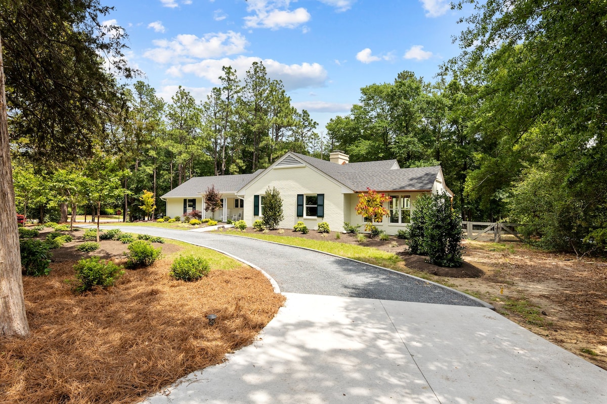 A ranch-style home is viewed from the driveway, surrounded by lush greenery and colorful landscaping. Large windows reflect the natural light, while a gentle curve in the driveway leads to a spacious front entrance. Towering trees provide a picturesque backdrop.