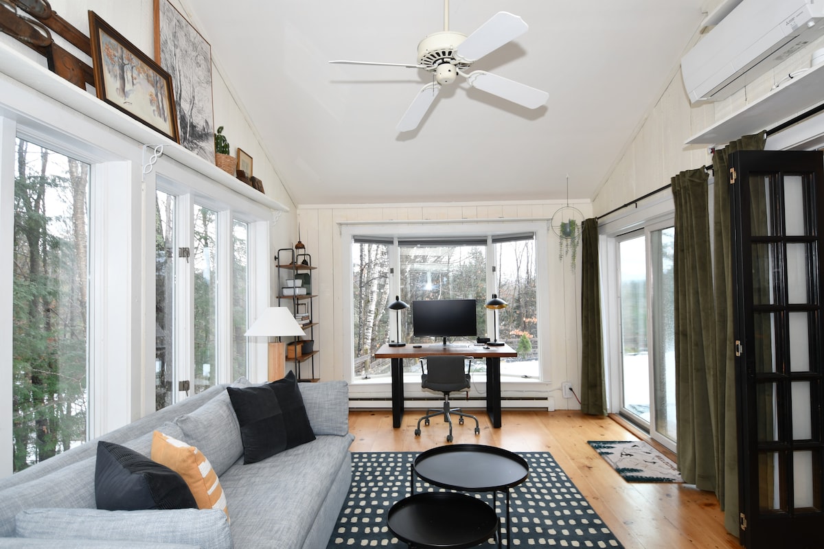 A bright and spacious living area is presented, featuring a comfortable gray sofa set against wooden flooring. A work-from-home station is positioned by large windows overlooking the outdoors. Natural light floods the room, complemented by ceiling fans and light-colored walls.
