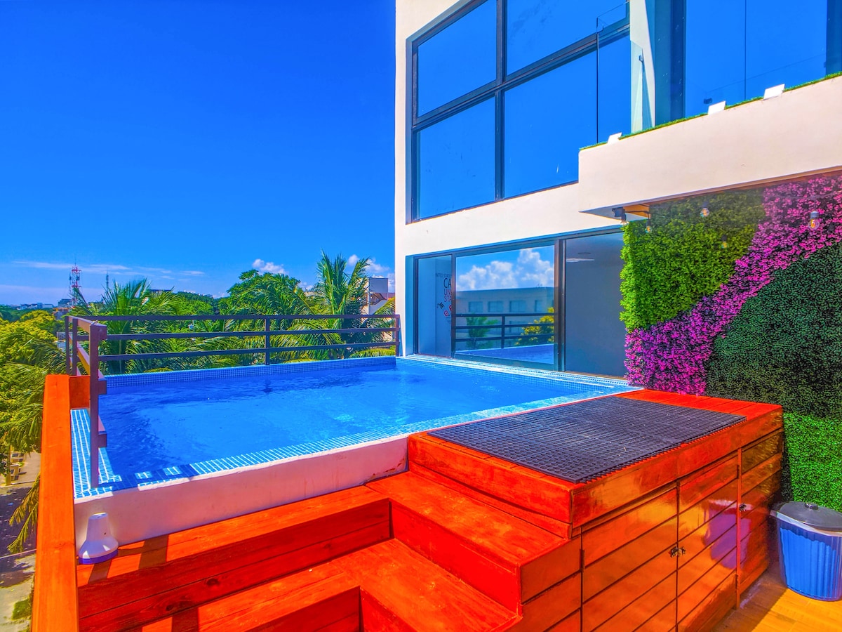 A rooftop pool is framed by colorful vertical greenery and wooden decking. Large glass windows provide a view of the surrounding landscape, while the clear blue water invites relaxation under the bright sun.