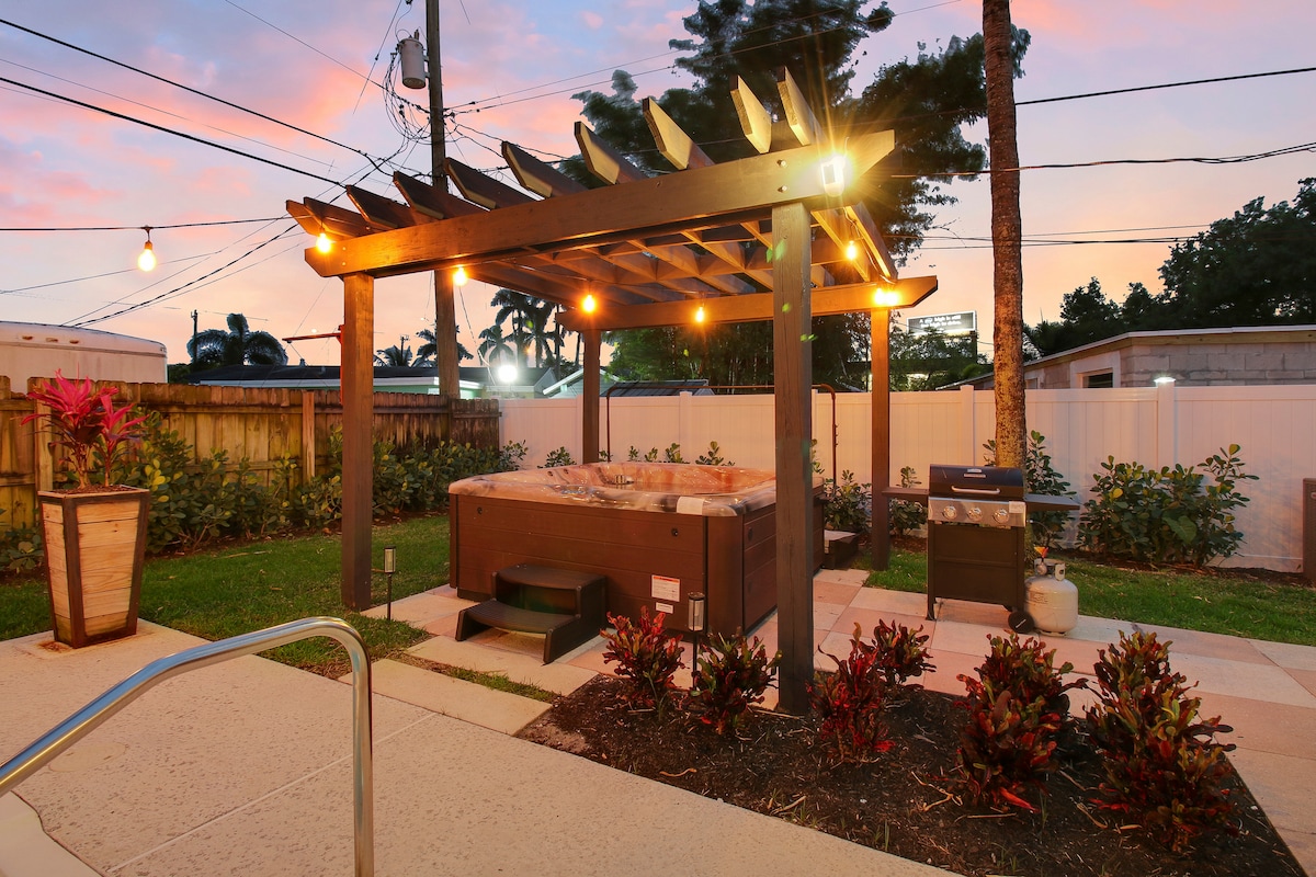 A private hot tub is nestled beneath a wooden pergola adorned with warm string lights. Surrounding greenery enhances the outdoor space, while a paved pathway leads to the hot tub area. A grill station is visible nearby, providing an inviting setting for relaxation.