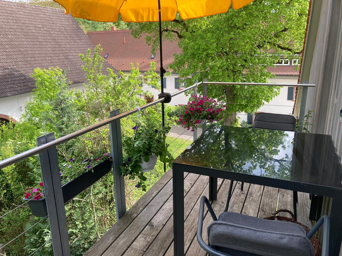 A balcony is shown with a sturdy table and chair arrangement under a yellow umbrella. Flower boxes with flowering plants adorn the railing, while lush greenery surrounds the area, providing a peaceful outdoor space for relaxation.
