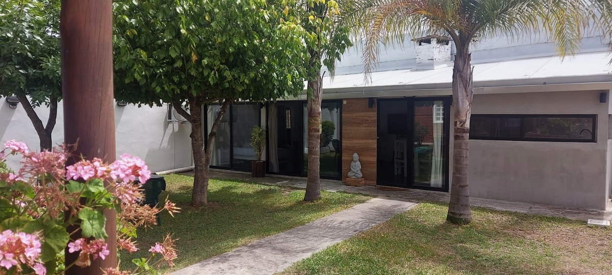 A serene garden area features lush greenery and flowering plants. The entrance to the apartment is framed by palm trees, with large glass doors partially revealing a modern interior. A decorative statue stands prominently along the pathway, enhancing the tranquil atmosphere.