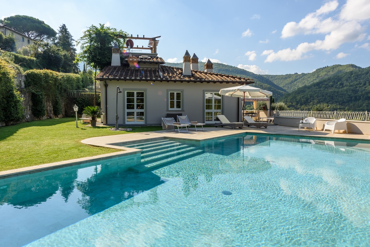 The image captures a luxurious villa with a private infinity pool, surrounded by lush green landscaping and hills. Sun loungers are positioned beside the pool, and the villa features a terrace with shaded seating. Large windows adorn the villa, providing ample natural light.