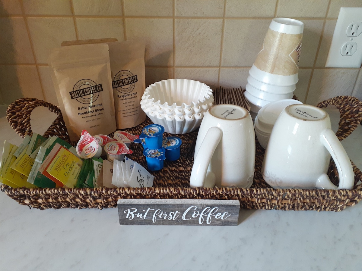 A woven tray displays multiple coffee supplies, including two white mugs, paper cups, a coffee filter holder, and assorted coffee creamers and tea bags. Two bags of ground coffee are also visible, along with a sign stating 'But first Coffee'.