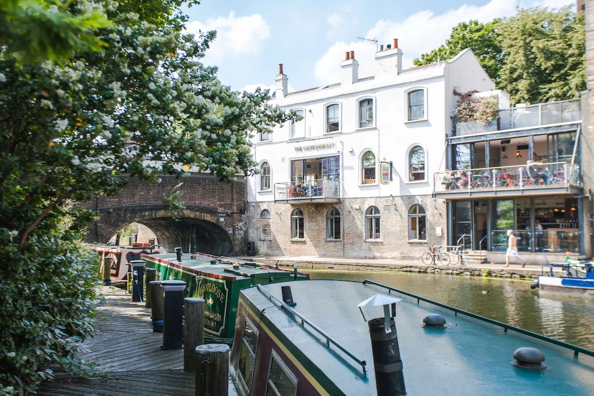 A scenic view along a canal features moored boats with bright green exteriors. A charming building stands adjacent to the water, showcasing multiple windows and balconies. Lush greenery and a stone bridge enhance the tranquil atmosphere of the area.